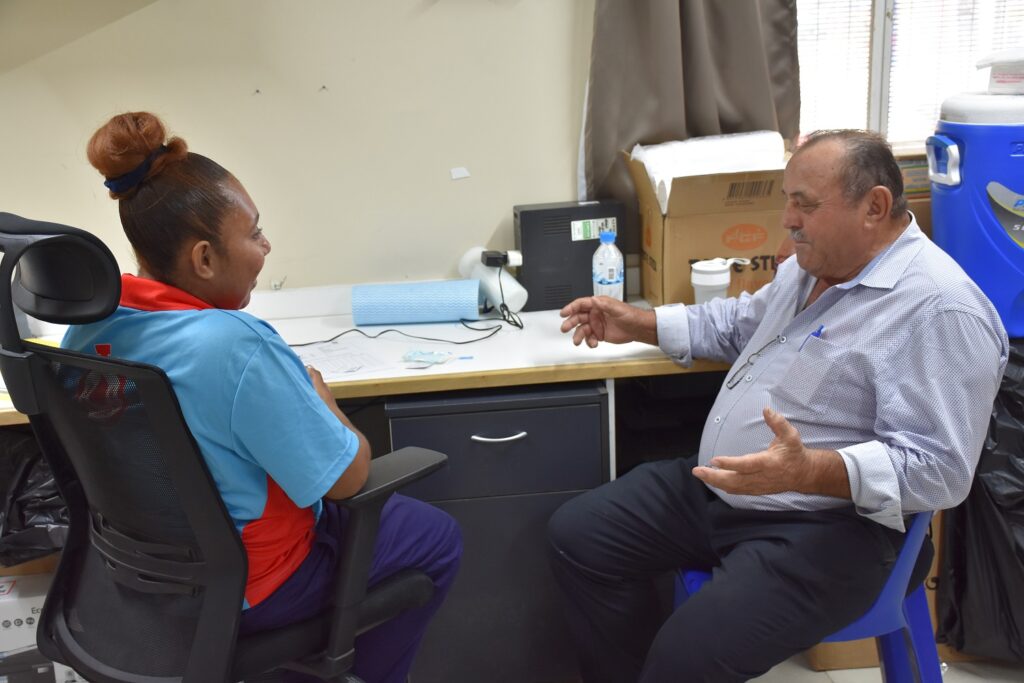 A woman in a blue shirt and a red collar sits in an office chair, engaged in conversation with a man in a light blue shirt seated on a blue plastic chair. They are discussing something over a desk cluttered with items.