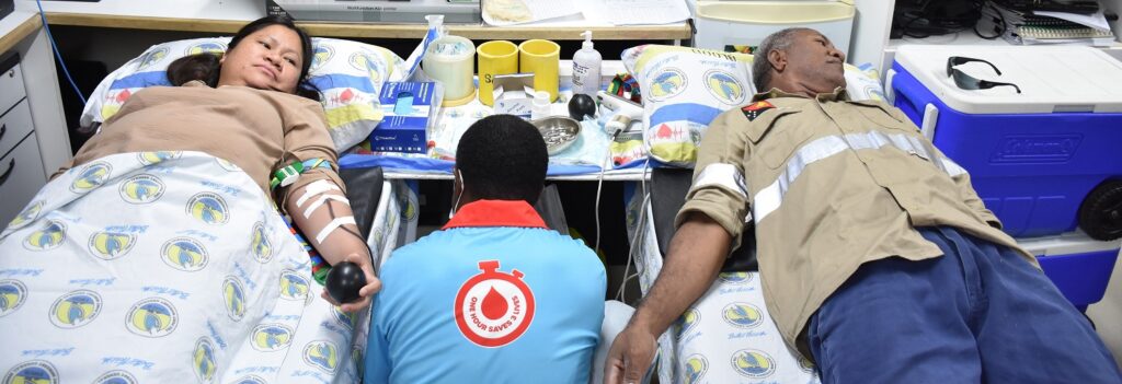 A blood donation session showing two donors lying on beds, with a medical professional attending to one of them. The setting includes medical equipment and supplies, and the donors are visibly engaged in the process.