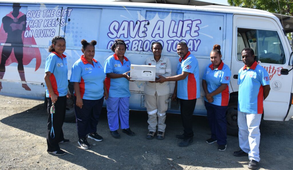 A group of eight people, including men and women, posing in front of a blood donation van that has slogans promoting blood donation. One person is handing over a box, possibly related to the blood donation process.